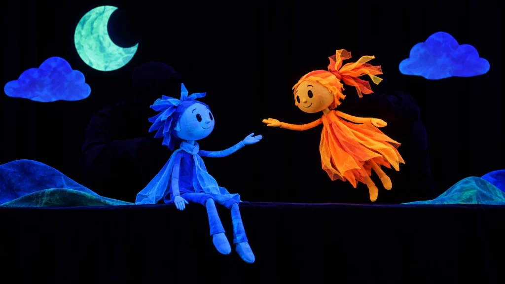 Escena de teatro negro con figuras iluminadas sobre un fondo oscuro.